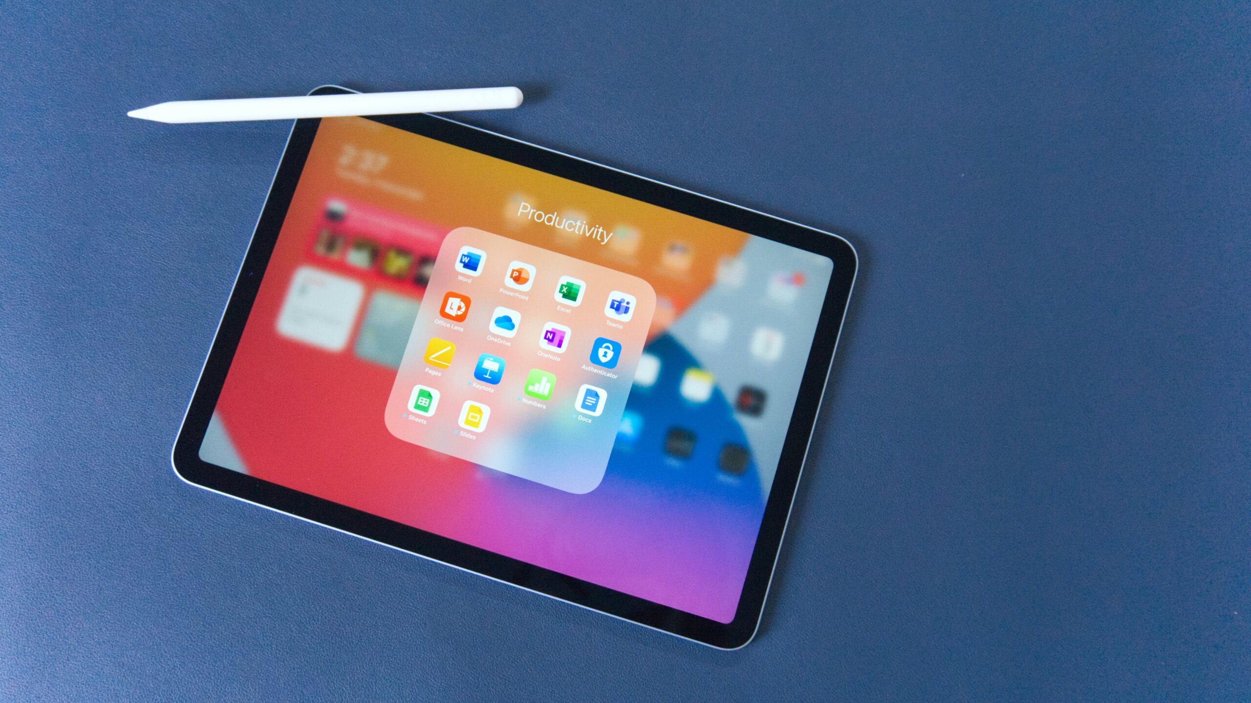 An iPad with a folder containing several apps open on the screen. The folder is labeled "Productivity" and the iPad is on a navy blue background. There is an apple pencil resting on the top left corner of the iPad. 